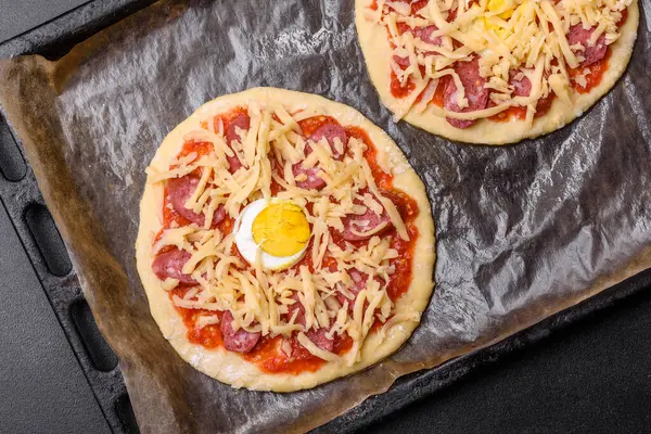 Homemade pizza with sausages, tomatoes, cheese, spices and herbs on a wooden cutting board against a dark concrete background