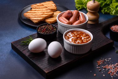Geleneksel İngiliz kahvaltısı yumurta, tost, sosis, fasulye, baharat ve yeşil seramik tabakta koyu beton arka planda otlar.