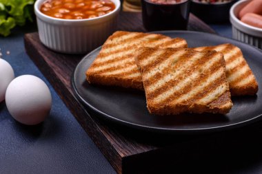 Geleneksel İngiliz kahvaltısı yumurta, tost, sosis, fasulye, baharat ve yeşil seramik tabakta koyu beton arka planda otlar.