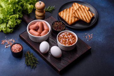 Geleneksel İngiliz kahvaltısı yumurta, tost, sosis, fasulye, baharat ve yeşil seramik tabakta koyu beton arka planda otlar.