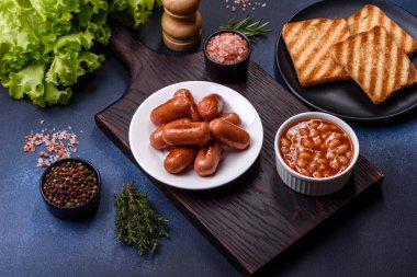 Geleneksel İngiliz kahvaltısı yumurta, tost, sosis, fasulye, baharat ve yeşil seramik tabakta koyu beton arka planda otlar.