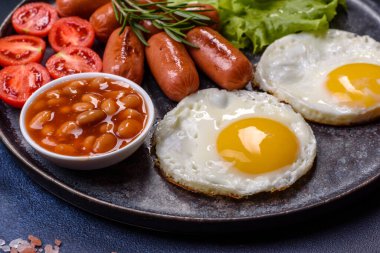 Geleneksel İngiliz kahvaltısı yumurta, tost, sosis, fasulye, baharat ve yeşil seramik tabakta koyu beton arka planda otlar.