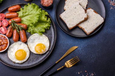 Geleneksel İngiliz kahvaltısı yumurta, tost, sosis, fasulye, baharat ve yeşil seramik tabakta koyu beton arka planda otlar.