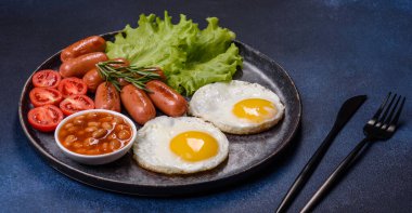 Geleneksel İngiliz kahvaltısı yumurta, tost, sosis, fasulye, baharat ve yeşil seramik tabakta koyu beton arka planda otlar.