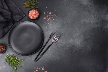İçinde baharatlar, otlar ve çatal bıçaklar olan koyu beton bir arka planda boş bir siyah tabak. Ev yemeği.