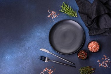 İçinde baharatlar, otlar ve çatal bıçaklar olan koyu beton bir arka planda boş bir siyah tabak. Ev yemeği.