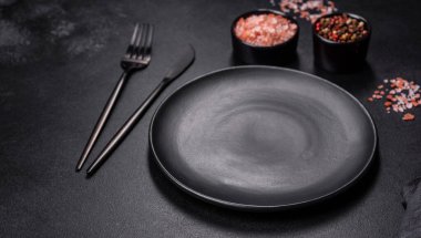 İçinde baharatlar, otlar ve çatal bıçaklar olan koyu beton bir arka planda boş bir siyah tabak. Ev yemeği.