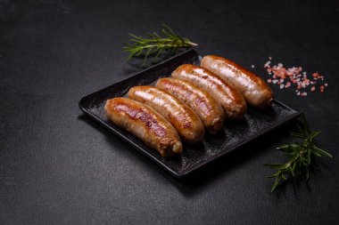 Lezzetli ızgara sosisler baharat ve otlarla siyah bir tabakta koyu beton arka planda.