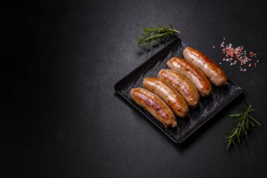 Lezzetli ızgara sosisler baharat ve otlarla siyah bir tabakta koyu beton arka planda.