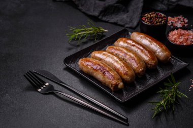 Lezzetli ızgara sosisler baharat ve otlarla siyah bir tabakta koyu beton arka planda.