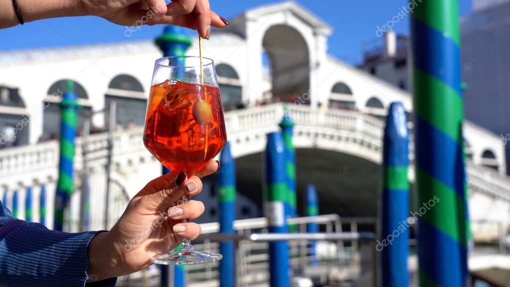 Girl holds in hand and drinks a glass of Spritz, in the city of love - aperitif, alcoholic drink Spritz Camapari or Aperol - typical drink of Venice made with  white wine, orange - Rialto bridge 