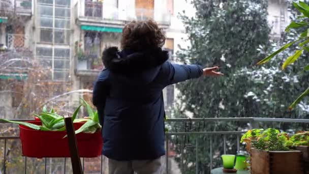 ヨーロッパ イタリア ミラノ 彼の家のバルコニーから雪 クリスマスの街の降雪 コロナウイルスCovid 19流行のために封鎖中に7歳の男の子 Omicronバリアント — ストック動画