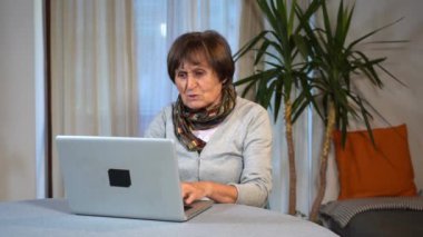 70 yaşında yaşlı bir kadın Covid-19 Coronavirus karantina evinde masada oturuyor dizüstü bilgisayarı kullanmayı, yazmayı ve internetten internet izlemeyi öğreniyor.
