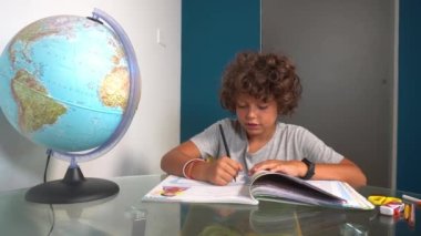 footage  of 8-year-old boy child does elementary school homework for summer at home during holidays - writing on textbook looking at globe during Covid-19 Coronavirus epidemic lockdown outbreak