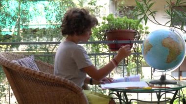 footage  of 8-year-old boy child does elementary school homework for summer at home during holidays - writing on textbook looking at globe during Covid-19 Coronavirus epidemic lockdown outbreak