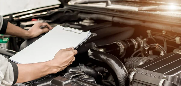 Automobile mechanic repairman checking a car engine with inspecting writing to the clipboard the checklist for repair machine, car service and maintenance.