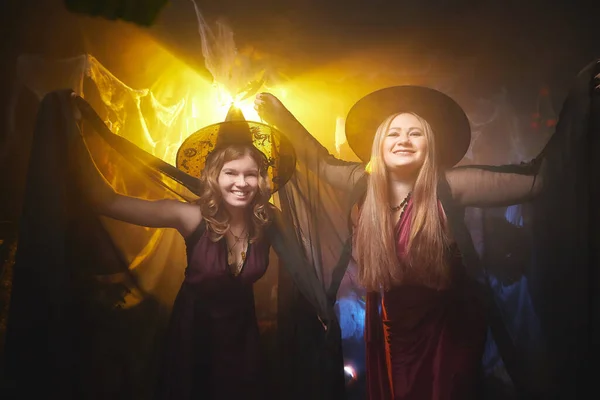 Two young women looking like witches are having fun on Halloween in a dark room with yellow light and smoke. Carnival concept and Halloween party
