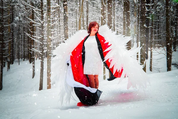 Woman looking as fairy angel with wings with white feathers in the forest with snow and snowdrifts on cold winter day. Model posing during photo shoot in cold park