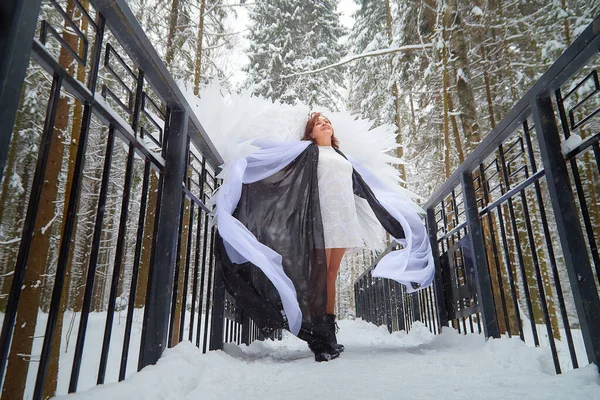 Woman looking as fairy angel with wings with white feathers in the forest with snow and snowdrifts on cold winter day. Model posing during photo shoot in cold park on the bridge