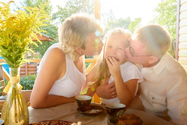 Little blonde girl, mother and father at a table on wooden terrace on nature or in the village and green landscape in the background on a summer or autumn sunny day. Concept happy family