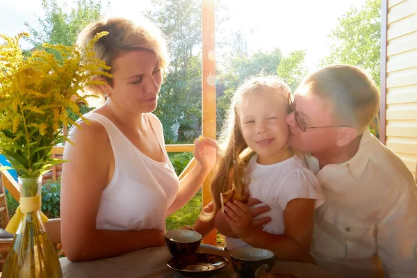 Little blonde girl, mother and father at a table on wooden terrace on nature or in the village and a green landscape in the background on a summer or autumn sunny day. Concept happy family
