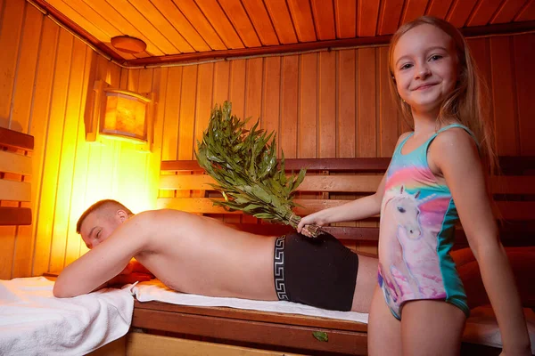 Man and young girl in a Russian bath. Bathhouse in Russia for a family with father and daughter having rest and fun together. The concept of healthy baby in the steam room with oak brooms