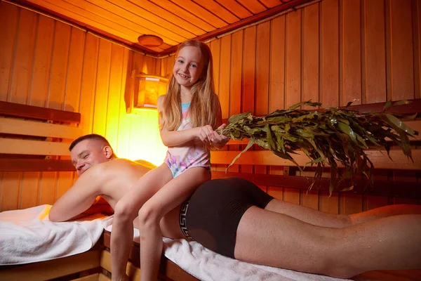 Man and young girl in a Russian bath. Bathhouse in Russia for a family with father and daughter having rest and fun together. The concept of healthy baby in the steam room with oak brooms