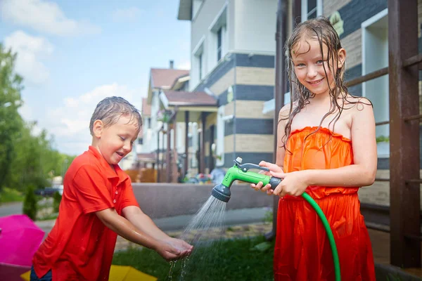 Children play with garden sprinkler. Brother and sister running and jumping. Summer outdoor water fun in backyard. Boy and girl play with hose watering grass. Kids run and splash on hot sunny day