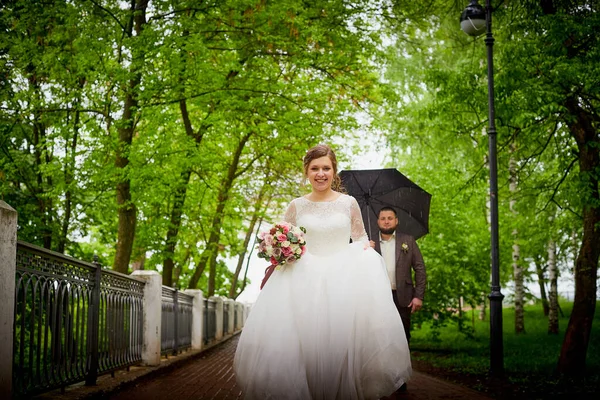 Wedding couple walks in green park with umbrella in rain in a green summer day