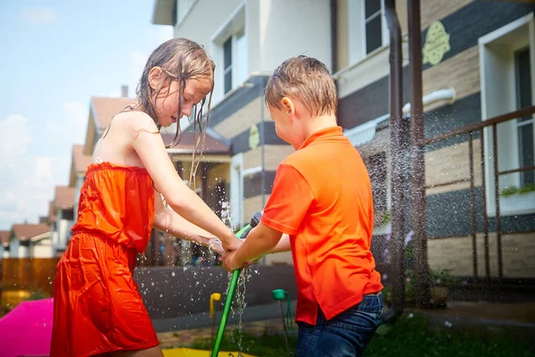 Children play with garden sprinkler. Brother and sister running and jumping. Summer outdoor water fun in backyard. Boy and girl play with hose watering grass. Kids run and splash on hot sunny day