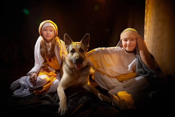 Little skinny girl and small boy in long white and yellow dress having fun with big dog. Young model sister and brother posing in dark studio in stylized Arabic east costume of Israel or Palestine