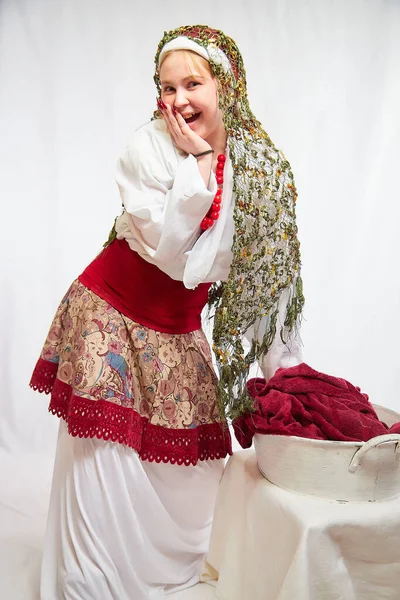 Beautiful smiling girl in stylized slavic red and white national costume washes the fabric in the basin on white background. Funny woman and model posing in studio as nice laundress