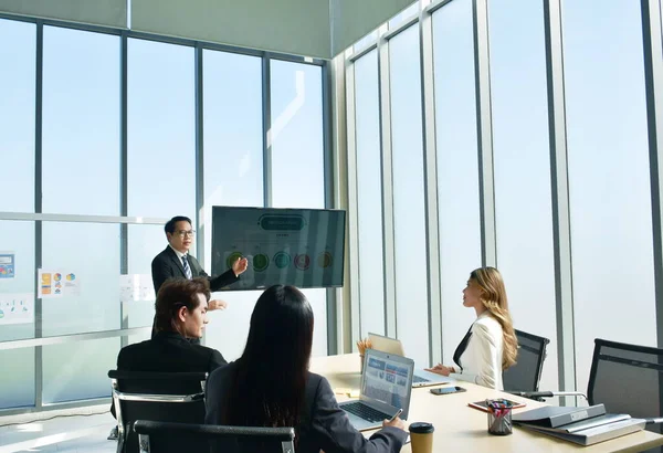 Asian business man presenting work on television screen with woman looking at computer laptop in office meeting room