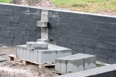 Temel duvarın içindeki nemli yalıtım, siyah asfalt bazlı karışım, paletlerin üzerinde beton bloklar olan bir inşaat alanı.