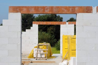 Reinforced brick lintels, floor to ceiling rough window openings and walls made of autoclaved aerated concrete, autoclaved aerated concrete blocks on pallets, a stairwell rough opening, blue sky and trees in the background