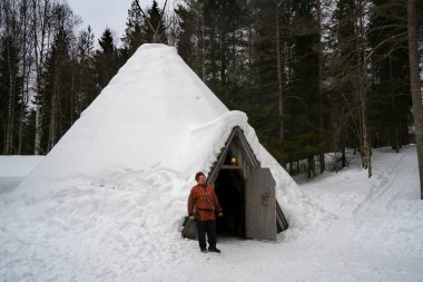Rovaniemi, Finlandiya - 18 Mart 2022: Geleneksel elbiseli bir sami adam, kar kaplı geleneksel sami ahşap evinin kapısında duruyor..