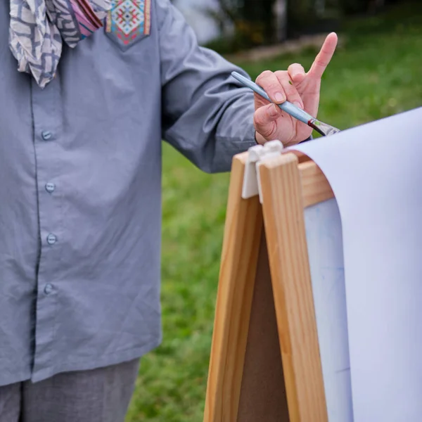 Happy woman draws on the easel with a brush and paints. Woman artist draws nature and trees on paper by the water on the river bank