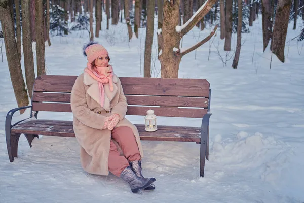 A happy woman is sitting on a bench with a lantern in her hands, a winter park with snow-covered trees
