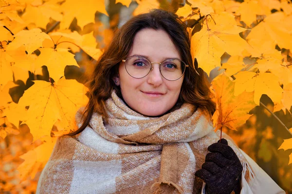 Portrait of an adult brunette woman with a scarf and glasses in autumn maple leaves