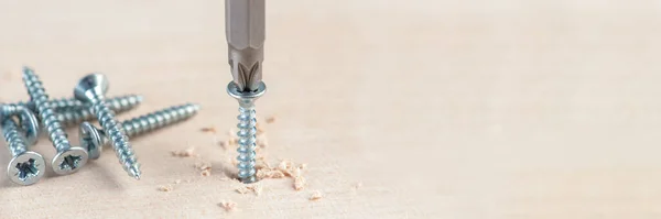 Screw the screw into the board. Furniture production, a screwdriver twists a self-tapping screw into a board. Several silver screws lie on the desktop — Stock Photo, Image