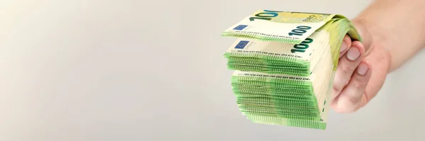 Stack of euro bills in hand. A large stack of 100 euro banknotes in a male hand on a uniform gray background. The concept of financial assistance, real estate purchase, loan or insurance payment