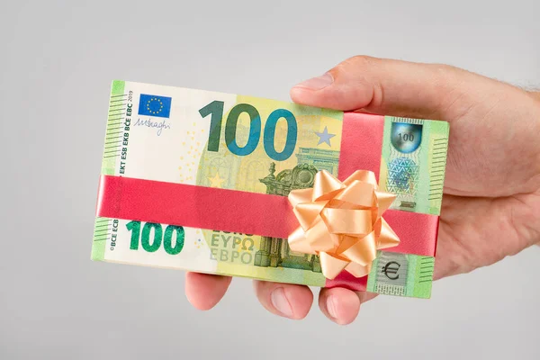 Gift of money. A pack of 100 euro bills in a mans hand. Money gift concept for birthday or christmas. Give euro with gift ribbon.