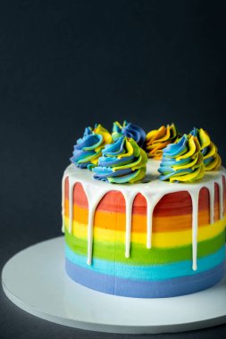 Koyu gri arka planda krem şantili gökkuşağı pastası. Çok renkli krem peynirli, kremalı ve beyaz çikolatalı pasta..