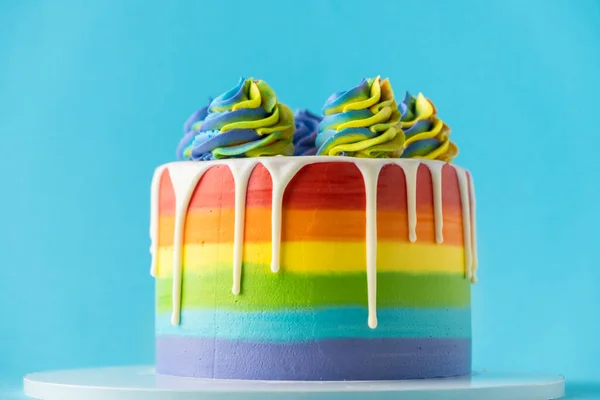 Rainbow cake with whipped cream top on the blue background. Birthday cake with multicolored cream cheese frosting and white chocolate drips.