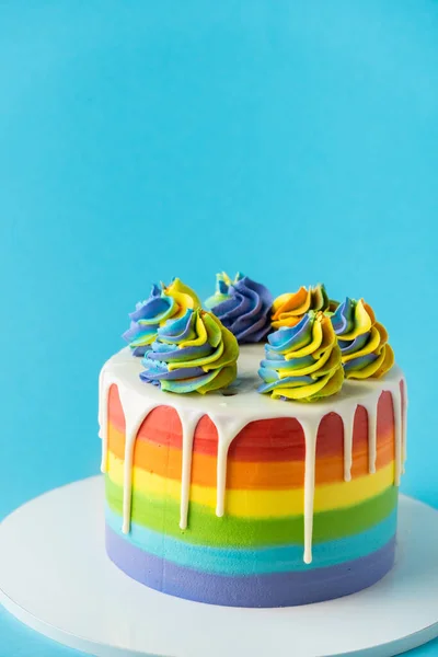 Rainbow cake with whipped cream top on the blue background. Birthday cake with multicolored cream cheese frosting and white chocolate drips.