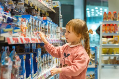 Küçük beyaz kız büyük bebek mağazasında yeni bir oyuncak seçiyor. Oyuncak dolu büyük raflar