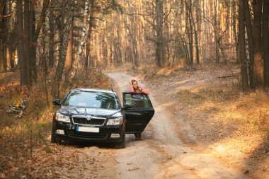 Kafası karışmış genç bir kadın arabanın yanında duruyor. Kapısı açık ve telefonda konuşuyor.