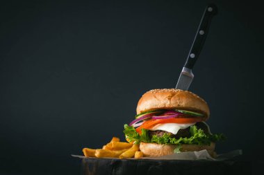 Büyük köfteli burger, peynirli, mor, soğanlı, salatalıklı, patates kızartmalı, ahşap, siyah, siyah arka planda.