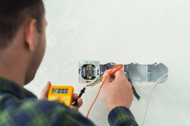 Elektrikçi beyaz bir duvara yeni bir soket takıyor. Kabloları kontrol etmek için çokölçer kullanan adam