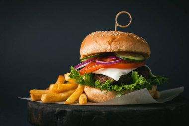 Büyük köfteli burger, peynirli, mor, soğanlı, salatalıklı, patates kızartmalı, ahşap, siyah, siyah arka planda.